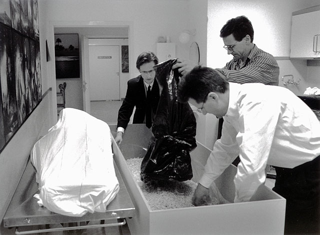 Two sons and a friend preparing the coffin with shavings, dead father wrapped in a white sheet