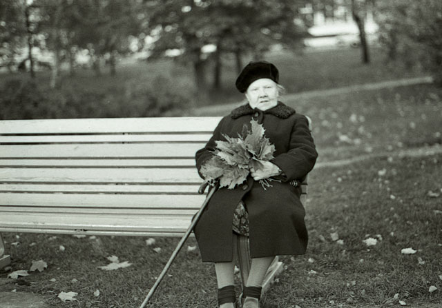 Old woman sitting on park bench in Helsinki Finland fall mapel leaves walking stick