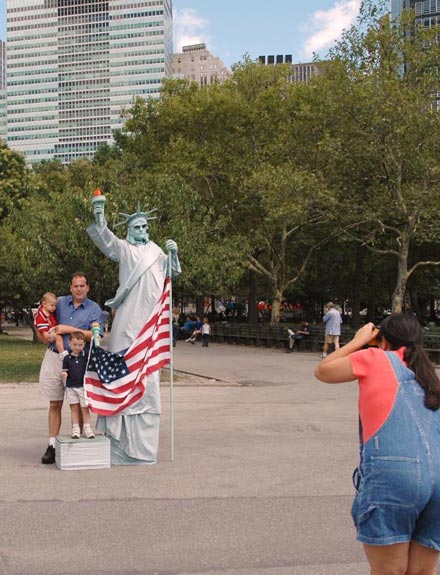 Wife photographing husbaned and two children in Battery park with a Statue of Liberty mime holding american flag
