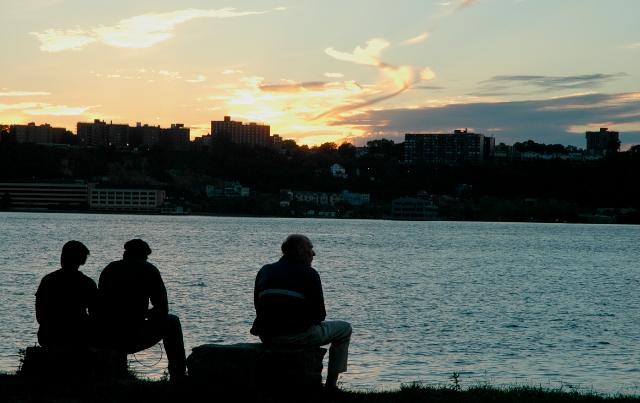 three men in silhouette sitting by Hudson river, sun setting behind buildings in New Jersey