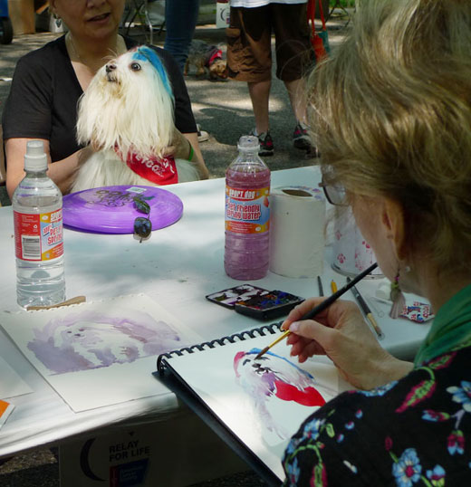 An artist painting a portrait of a Lhasa Apso with blue and red in his hair, Riverside Park a dog event